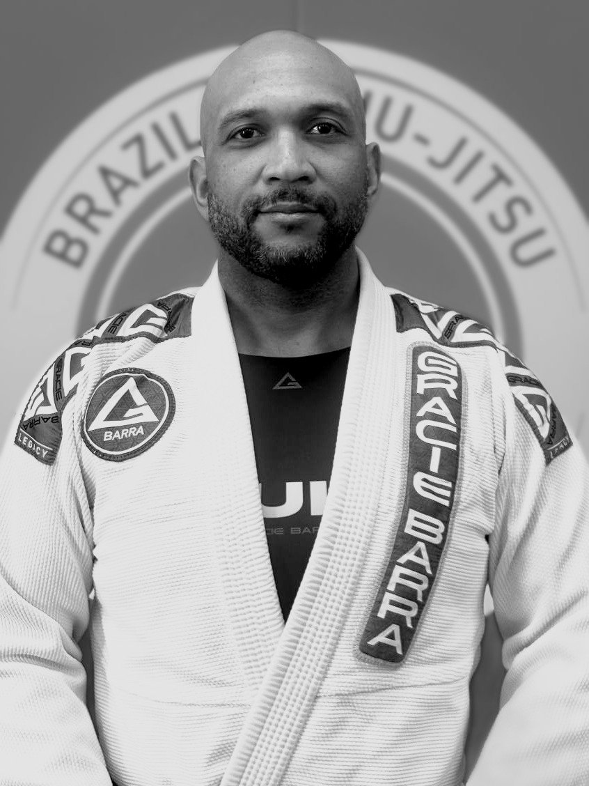 Coach Keith Gumbs, brown belt instructor at Gracie Barra Seminole, standing in his gi in front of a Gracie Barra Brazilian Jiu-Jitsu backdrop
